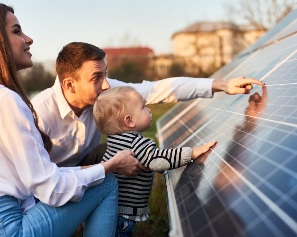 Side close-up shot of a young modern family with a little son getting acquainted with solar panel on a sunny day, green alternative energy concept